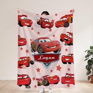 May include: A light pink blanket with various illustrations of Lightning McQueen from the film Cars. The blanket includes red stars and a nameplate that reads "Logan".