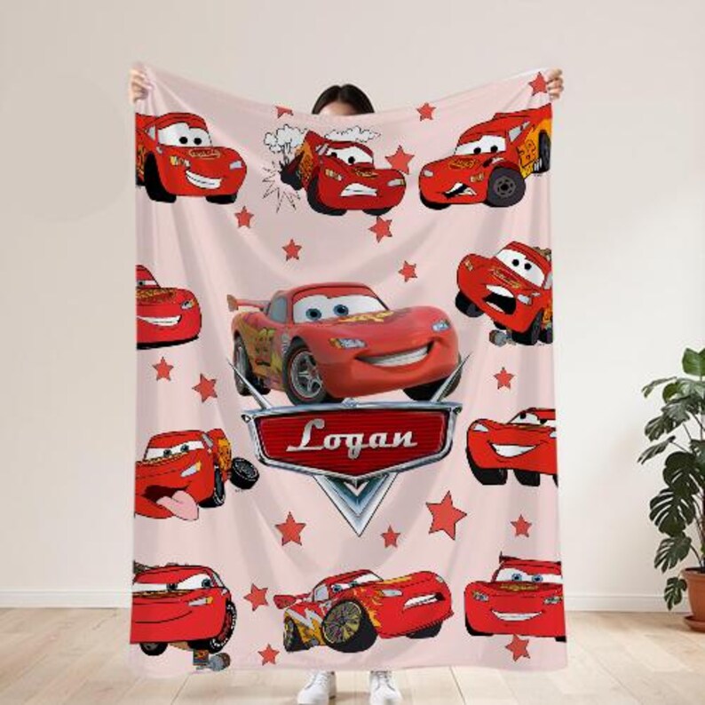 May include: A light pink blanket with various illustrations of Lightning McQueen from the film Cars. The blanket includes red stars and a nameplate that reads "Logan".