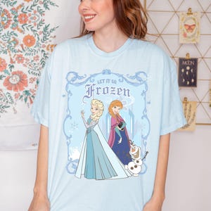 May include: Light blue t-shirt featuring Elsa, Anna, and Olaf from the movie Frozen. The graphic includes the text "Let It Go" and "Frozen" in a decorative frame. The characters are depicted in their signature outfits.