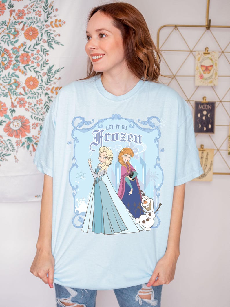 May include: Light blue t-shirt featuring Elsa, Anna, and Olaf from the movie Frozen. The graphic includes the text "Let It Go" and "Frozen" in a decorative frame. The characters are depicted in their signature outfits.