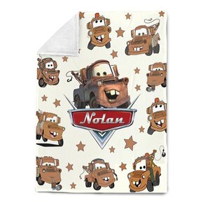 May include: A cream-colored blanket featuring a repeating pattern of the character Mater from the movie Cars, along with brown stars. The blanket has the name "Nolan" in a red and silver emblem.