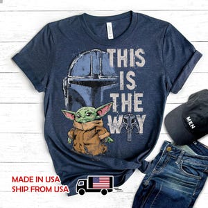 May include: A navy blue short-sleeve t-shirt featuring a graphic of a green creature in a tan robe, a blue helmet, and a skull icon. The text "THIS IS THE WAY" is printed in white. The shirt has a crew neckline and a heathered fabric texture.