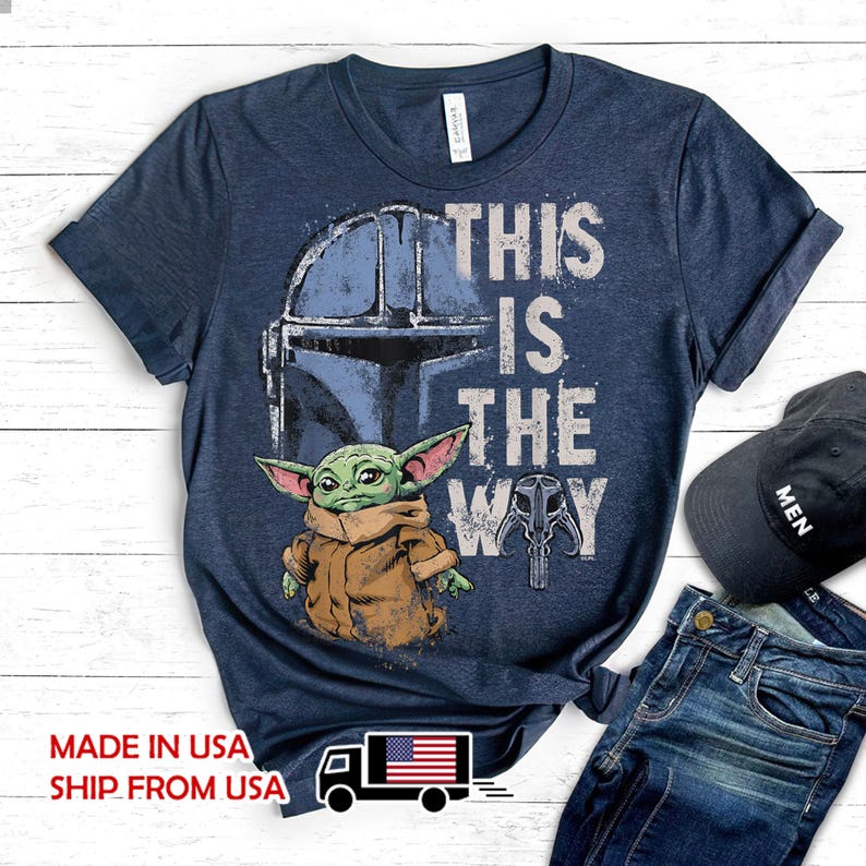 May include: A navy blue short-sleeve t-shirt featuring a graphic of a green creature in a tan robe, a blue helmet, and a skull icon. The text "THIS IS THE WAY" is printed in white. The shirt has a crew neckline and a heathered fabric texture.