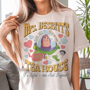 Op de afbeelding: Cr&egrave;mekleurig T-shirt met een cartoonafbeelding van Buzz Lightyear uit Toy Story. Het ontwerp bevat de tekst "Mrs. Nesbitt's Tea House" met harten en een theepot. De zin "To Infini-tea - tea And Beyond" is ook aanwezig.