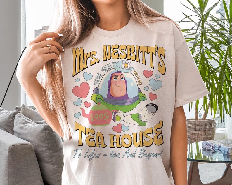 Op de afbeelding: Cr&egrave;mekleurig T-shirt met een cartoonafbeelding van Buzz Lightyear uit Toy Story. Het ontwerp bevat de tekst "Mrs. Nesbitt's Tea House" met harten en een theepot. De zin "To Infini-tea - tea And Beyond" is ook aanwezig.