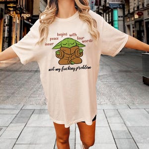 Może przedstawiać: Kremowy t-shirt z kr&oacute;tkim rękawem z grafiką przedstawiającą zielone stworzenie w brązowej szacie z zamkniętymi oczami. Tekst brzmi: peace begins with inner four words: not my fucking problem. Koszulka posiada okrągły dekolt i luźny kr&oacute;j.