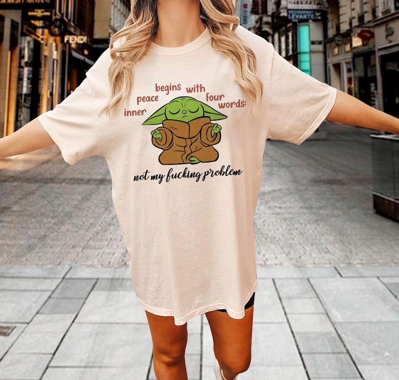 Może przedstawiać: Kremowy t-shirt z kr&oacute;tkim rękawem z grafiką przedstawiającą zielone stworzenie w brązowej szacie z zamkniętymi oczami. Tekst brzmi: peace begins with inner four words: not my fucking problem. Koszulka posiada okrągły dekolt i luźny kr&oacute;j.