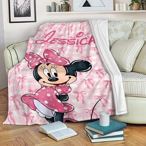 May include: A pink and white blanket featuring a Minnie Mouse design. The blanket has a repeating pattern of bows and the name "Jessica" in pink script. Minnie Mouse is wearing a pink polka dot dress.
