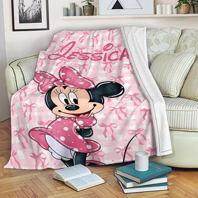 May include: A pink and white blanket featuring a Minnie Mouse design. The blanket has a repeating pattern of bows and the name "Jessica" in pink script. Minnie Mouse is wearing a pink polka dot dress.