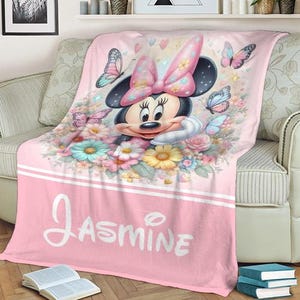 Op de afbeelding: Roze deken met Minnie Mouse met een grote roze strik, omringd door bloemen en vlinders. De naam "Jasmine" staat in witte letters onderaan. De deken ligt over een beige bank.