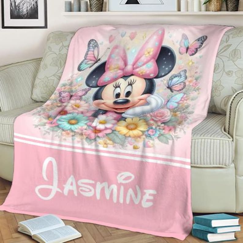 Op de afbeelding: Roze deken met Minnie Mouse met een grote roze strik, omringd door bloemen en vlinders. De naam "Jasmine" staat in witte letters onderaan. De deken ligt over een beige bank.