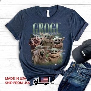 May include: A dark blue heathered short-sleeve t-shirt featuring a graphic print of five Grogu characters in tan robes. The text GROGU is printed in a large, light green font above the characters, which are surrounded by subtle lightning effects.