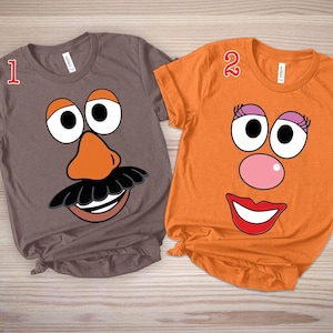 Op de afbeelding: Twee T-shirts met korte mouwen, één grijs en één oranje, elk met een cartoongezicht. Het grijze shirt heeft een gezicht met een bruine neus, een zwarte snor en een lachende mond. Het oranje shirt heeft een roze neus, rode lippen en wimpers.