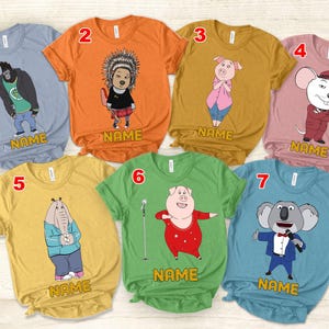 May include: Seven colourful t-shirts featuring cartoon characters from the film "Sing." Each shirt has a different character design, including a gorilla, a porcupine, a pig, a mouse, an elephant, another pig, and a koala. The word "NAME" is printed on each shirt.