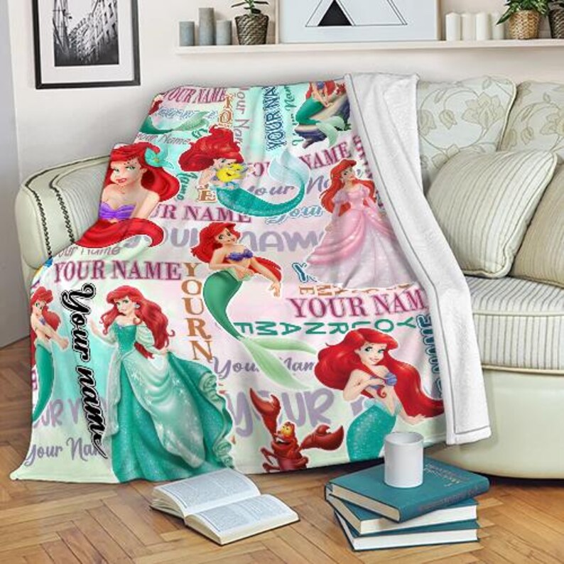 May include: A soft, light-colored blanket featuring multiple illustrations of a mermaid with red hair and a green tail. The blanket has the text "YOUR NAME" in various fonts and colors, with additional cartoon characters.