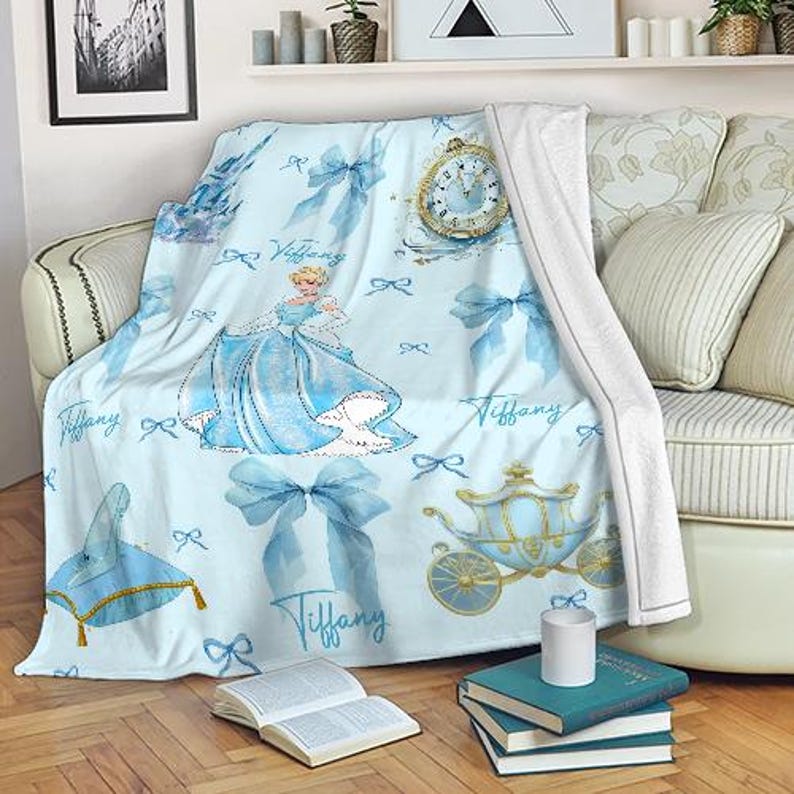 K&ouml;nnte beinhalten: Hellblaue Decke mit einer Prinzessin in blauem Kleid, Schleifen, einer Kutsche und einer Uhr. Das Wort "Tiffany" ist auf die Decke geschrieben. Die Decke ist &uuml;ber einem Sofa mit wei&szlig;en Kissen drapiert.