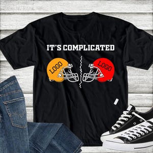 Może przedstawiać: Czarny t-shirt z napisem "IT'S COMPLICATED" nad dwoma kaskami do futbolu amerykańskiego, jednym pomarańczowym i jednym czerwonym. Na każdym kasku znajduje się słowo "LOGO". Koszulka jest zestawiona z niebieskimi dżinsami i czarnymi trampkami.