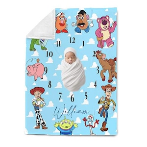 May include: A baby blanket featuring a light blue background with cartoon characters and a clock design. The blanket has a white border and the name "William" in cursive. The blanket is decorated with characters from the movie Toy Story.