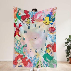 May include: A rectangular blanket featuring a colorful underwater scene with cartoon mermaids, fish, and a crab. The design includes a clock face with blue numerals and aquatic plants. The blanket is held up by a person.