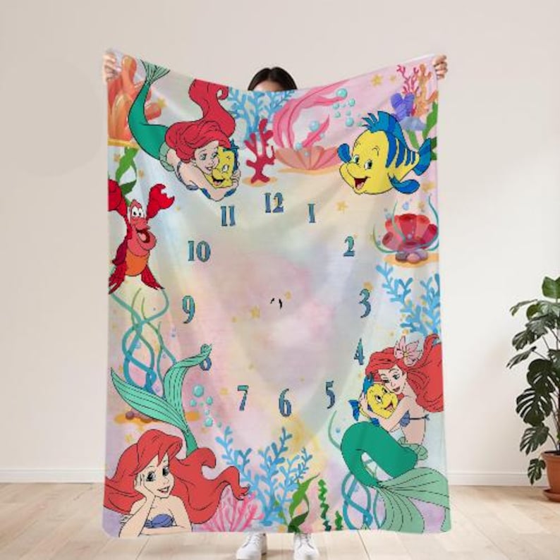 May include: A rectangular blanket featuring a colorful underwater scene with cartoon mermaids, fish, and a crab. The design includes a clock face with blue numerals and aquatic plants. The blanket is held up by a person.