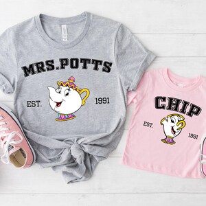 Puede incluir: Dos camisetas, una gris y otra rosa, con personajes de dibujos animados. La camiseta gris dice "MRS. POTTS" con un gráfico de tetera y "EST. 1991". La camiseta rosa dice "CHIP" con un gráfico de taza y "EST. 1991". Zapatillas rosas en la imagen.