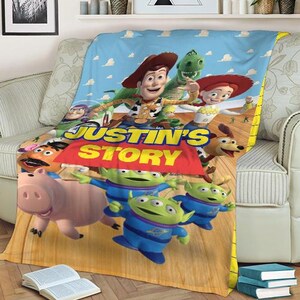May include: A plush blanket featuring characters from the animated film Toy Story. The blanket displays Woody, Rex, Jessie, and other characters against a blue sky background. The text "JUSTIN'S STORY" is prominently displayed.