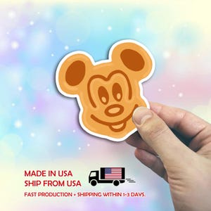 May include: A tan sticker in the shape of Mickey Mouse's face, held by a hand. The sticker has a white border and a smiling face. The background is a soft blend of blue, pink, and white with bokeh effects.