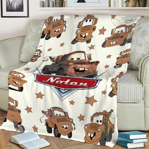 May include: A cream-colored blanket featuring a repeating pattern of a cartoon tow truck character and brown stars. The blanket has a large image of the character with the name "Nolan" on a red and silver emblem.