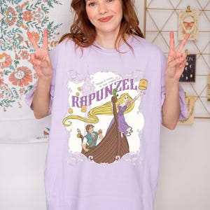 May include: A light purple t-shirt featuring a graphic of Rapunzel from the Disney movie Tangled. The design includes Rapunzel in a boat with a lantern, and the text "Rapunzel" and "There's magic in the world."