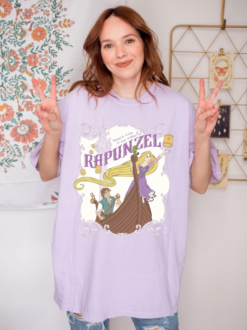 May include: A light purple t-shirt featuring a graphic of Rapunzel from the Disney movie Tangled. The design includes Rapunzel in a boat with a lantern, and the text "Rapunzel" and "There's magic in the world."