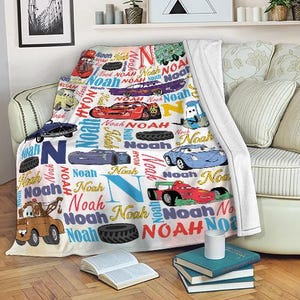 May include: A white blanket featuring colorful illustrations of race cars and the name "Noah" in various fonts and colors. The blanket is draped over a sofa, with a stack of books and a mug in the foreground.
