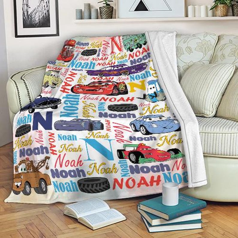 May include: A white blanket featuring colorful illustrations of race cars and the name "Noah" in various fonts and colors. The blanket is draped over a sofa, with a stack of books and a mug in the foreground.