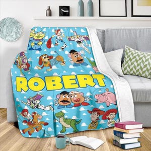 K&ouml;nnte beinhalten: Eine hellblaue Decke mit Figuren aus dem Film Toy Story, mit dem Namen "ROBERT" in gro&szlig;en gelben Buchstaben. Die Decke hat eine wei&szlig;e R&uuml;ckseite und ist &uuml;ber ein graues Sofa drapiert. Die Decke ist mit Zeichentrickfiguren verziert.