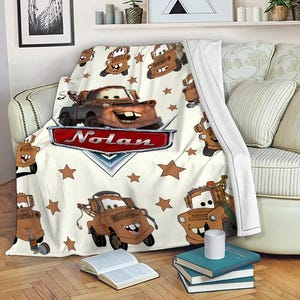 May include: A cream-colored blanket featuring a cartoon tow truck character with a smiling face. The blanket has a red nameplate that says "Nolan" and brown star shapes. The blanket is draped over a sofa.