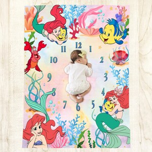 May include: A baby milestone blanket featuring illustrations of Ariel, Flounder, and Sebastian from The Little Mermaid. The blanket has a watercolor background with a clock design and numbers. A baby is lying on the blanket.