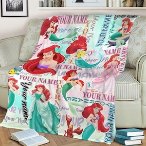 May include: A soft, fleece blanket featuring multiple illustrations of a mermaid with red hair and a teal tail. The blanket has the words "YOUR NAME" in a repeating pattern. The mermaid is wearing a purple top and a teal tail.
