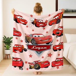 May include: A light pink blanket featuring multiple images of a red race car with a smiling face. The blanket has red star shapes and the name "Logan" on a silver emblem.