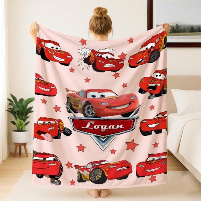 May include: A light pink blanket featuring multiple images of a red race car with a smiling face. The blanket has red star shapes and the name "Logan" on a silver emblem.