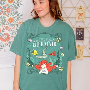 May include: Green t-shirt featuring a graphic of Ariel from The Little Mermaid. The design includes Ariel with red hair, a mermaid tail, and other characters. The text "The Little Mermaid" is displayed above the graphic.