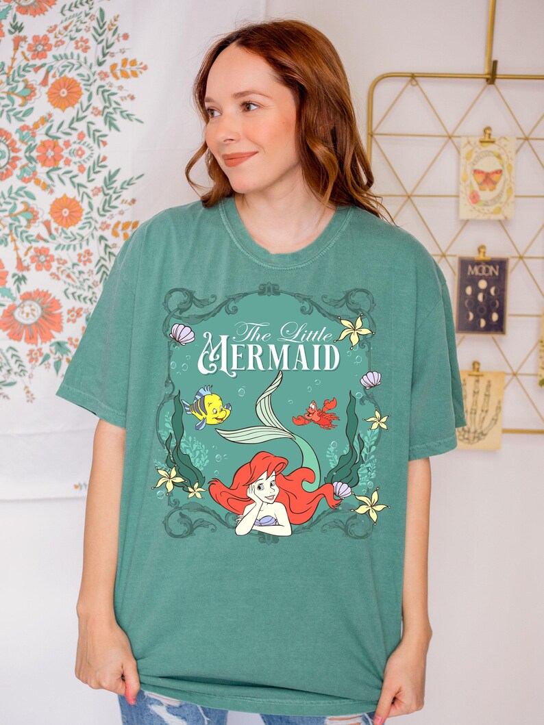 May include: Green t-shirt featuring a graphic of Ariel from The Little Mermaid. The design includes Ariel with red hair, a mermaid tail, and other characters. The text "The Little Mermaid" is displayed above the graphic.