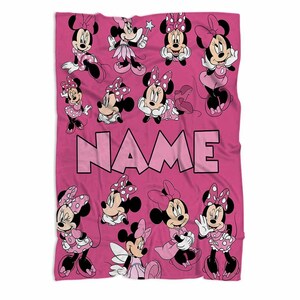 May include: A pink blanket featuring multiple illustrations of Minnie Mouse in various poses. The blanket has the word "NAME" in large pink letters across the center. Minnie Mouse is wearing pink dresses and bows with white polka dots.