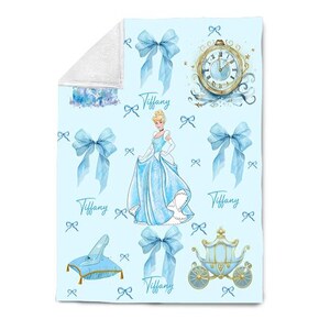 K&ouml;nnte beinhalten: Eine hellblaue Decke mit einem Aquarelldesign mit Aschenputtel, Schleifen, einer Kutsche, einer Uhr und einem Glasschuh. Der Name "Tiffany" ist kursiv geschrieben. Die Decke hat eine wei&szlig;e, flauschige R&uuml;ckseite.
