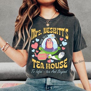 Op de afbeelding: Donkergrijs T-shirt met de tekst "Mrs. Nesbitt's Tea House" en een cartoonfiguur. Het ontwerp bevat harten, een theepot en de zin "To Infini - tea And Beyond."
