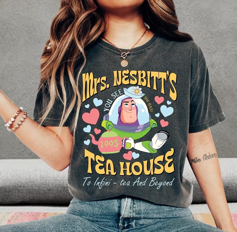 Op de afbeelding: Donkergrijs T-shirt met de tekst "Mrs. Nesbitt's Tea House" en een cartoonfiguur. Het ontwerp bevat harten, een theepot en de zin "To Infini - tea And Beyond."