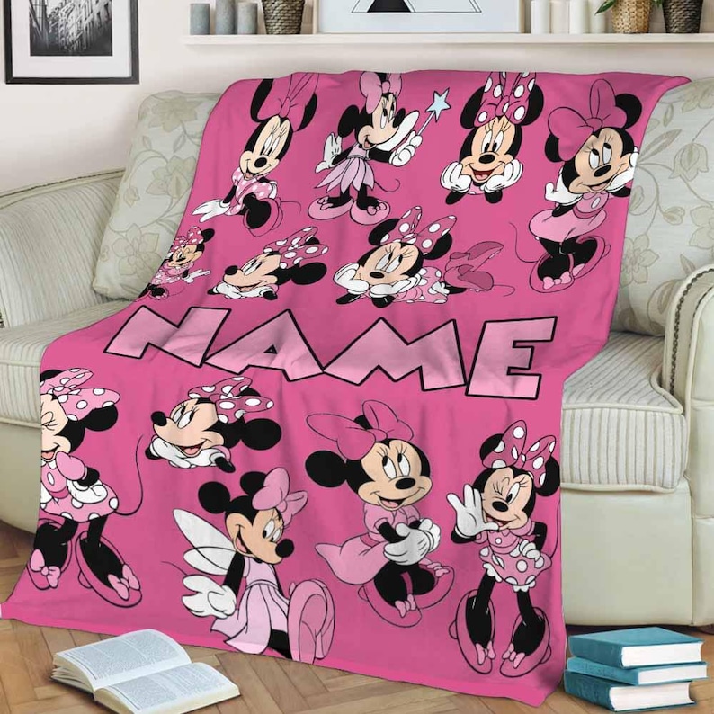 May include: Pink fleece blanket featuring multiple illustrations of Minnie Mouse in various poses. The blanket has the word "NAME" in large, light pink letters across the center. The blanket is draped over a light-colored sofa.