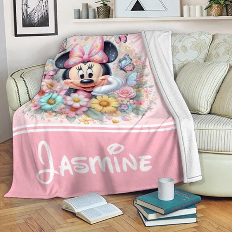Op de afbeelding: Roze deken met Minnie Mouse omringd door bloemen en vlinders. De deken heeft de naam "Jasmine" in witte letters over de bodem. Het ontwerp heeft een witte rand.