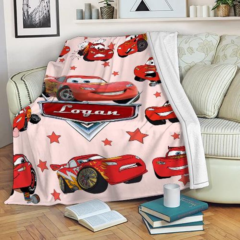 May include: A light pink blanket with illustrations of red race cars and red stars. The blanket has the name "Logan" on a silver emblem. The blanket is draped over a sofa.