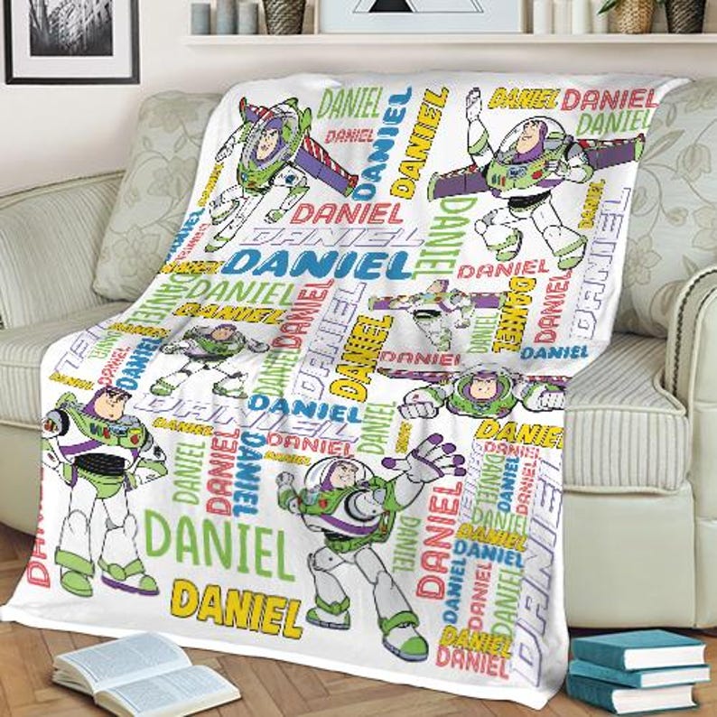 May include: White blanket featuring multiple images of Buzz Lightyear in various poses, with the name "DANIEL" repeated in colorful text. The blanket is draped over a sofa.