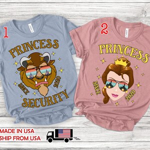 Pode incluir: Duas camisetas de manga curta. Uma é azul clara com um gráfico da Fera, a outra é rosa com um gráfico da Bela. Ambas as camisas têm a palavra "PRINCESS" acima do rosto do personagem e "SINCE 2020 SECURITY" abaixo.