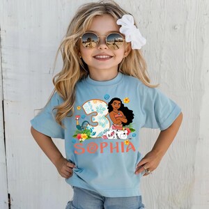 Puede incluir: Una camiseta azul claro con un gr&aacute;fico colorido que presenta un personaje de dibujos animados, el n&uacute;mero 3 y el nombre "Sophia" en coral. El gr&aacute;fico incluye elementos florales y animales de dibujos animados. La camiseta es para un ni&ntilde;o.